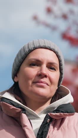 mujer de negocios con chaqueta rosa y gorra gris, mirando pensativa con la cabeza inclinada, al aire libre en otoño, posando con árboles de colores borrosos y casa en el fondo bajo un cielo despejado