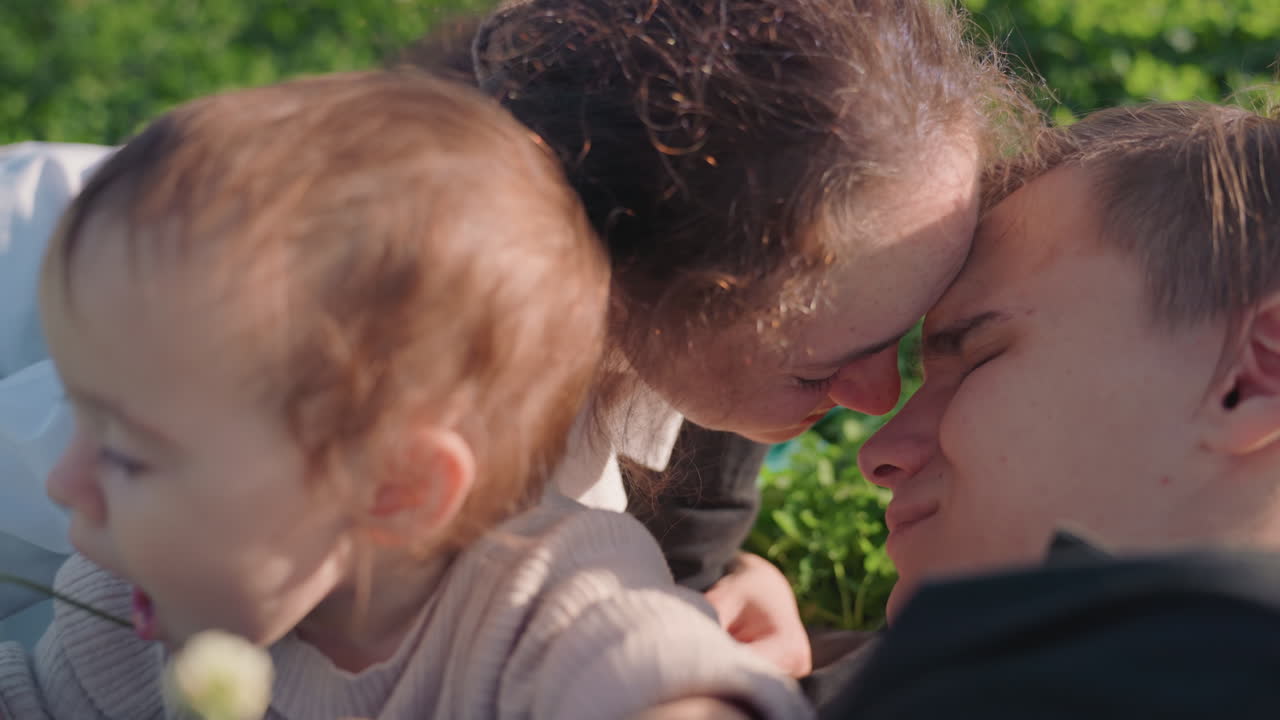 Familie spielt im grünen Gras, Mutter und Vater erkunden mit Kleinkind Klee, Baby greift nach Blume, Eltern küssen sich, helles Sonnenlicht, authentischer Lifestyle-Moment, fürsorgliche Familienbindung.