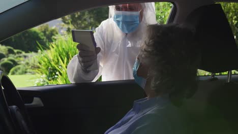 Caucasian-senior-woman-in-car-wearing-face-mask-having-temperature-taken-by-man-in-ppe-suit