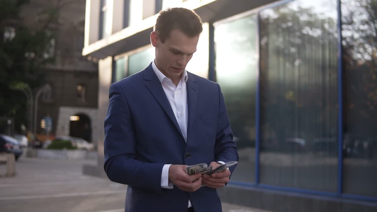 красивый кавказский бизнесмен в синем твердом костюме и белой рубашке, стоящий на улице и считающий американские банкноты