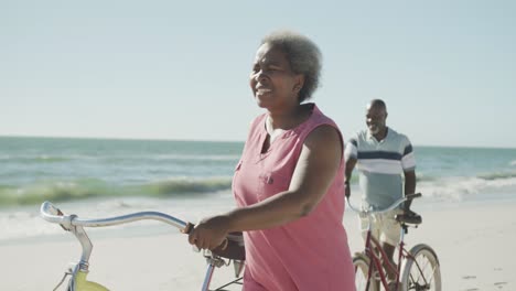 Feliz-Pareja-Afroamericana-Caminando-Con-Bicicletas-En-La-Playa,-En-Cámara-Lenta
