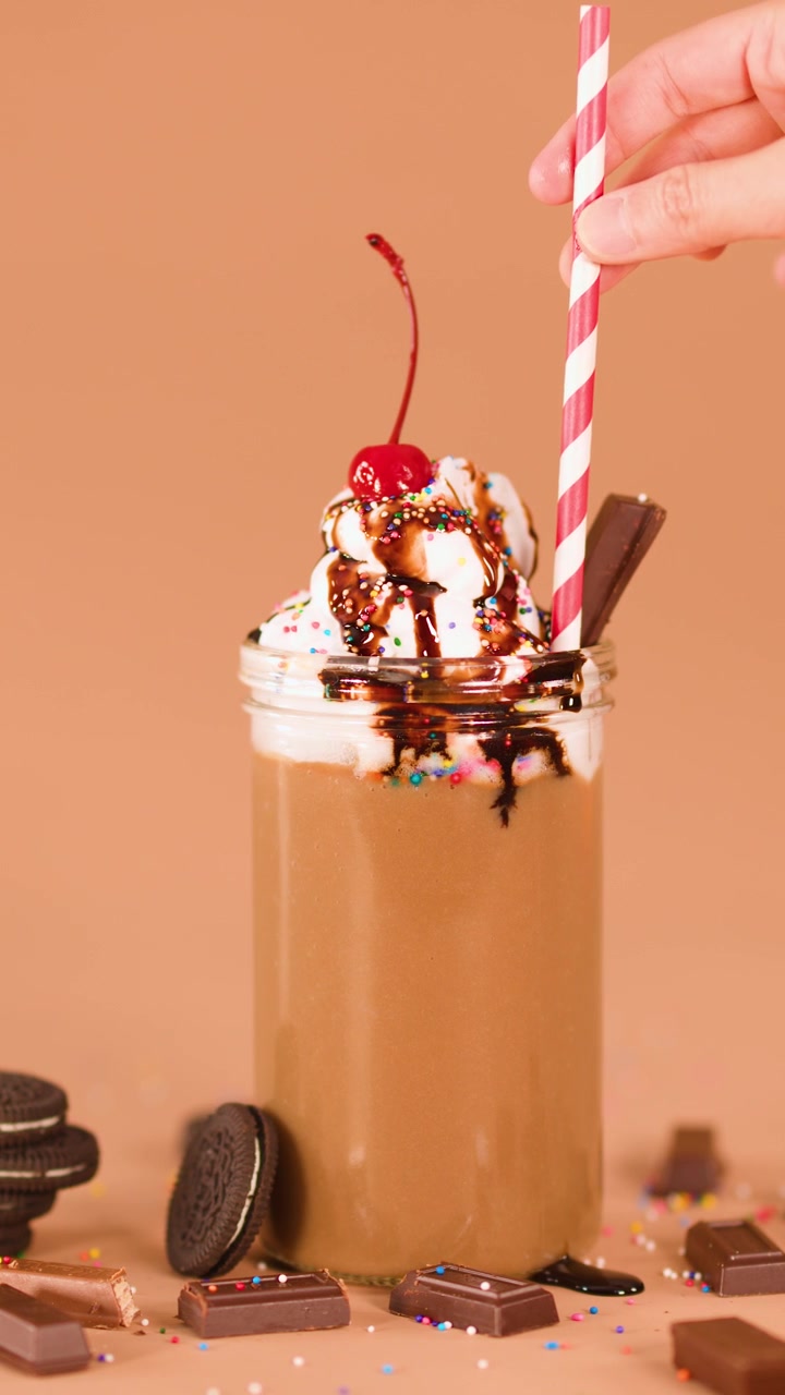Hand places striped straw in chocolate milkshake topped with whipped cream, cherry, and chocolate syrup