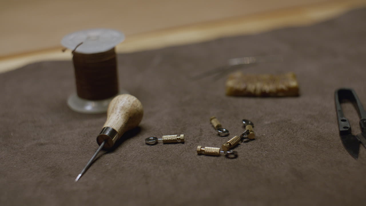 A close-up of leatherworking tools including an awl, thread spool, metal clasps, and cutting tools laid out on a brown suede work surface.