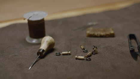 a close-up of leatherworking tools including an awl, thread spool, metal clasps, and cutting tools laid out on a brown suede work surface.