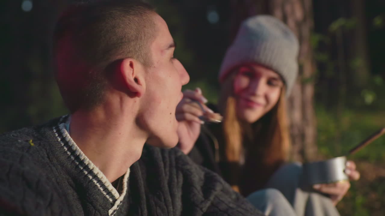 romantic campfire scene at dusk with woman feeding partner from can beside tent glow under forest night canopy intimate mood couple share meal laughter connection warmth joy outdoors