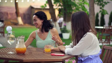 two women sitting at the outdoors cafe - talking, sharing news, brunette girl putting laptop on table and promoting something