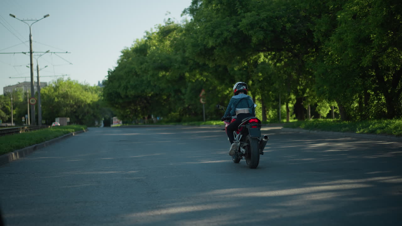 una dama monta una bicicleta eléctrica a lo largo de una carretera alineada con árboles verdes y postes eléctricos, con su camisa ondeando en el viento, exponiendo su espalda, con un coche visto delante