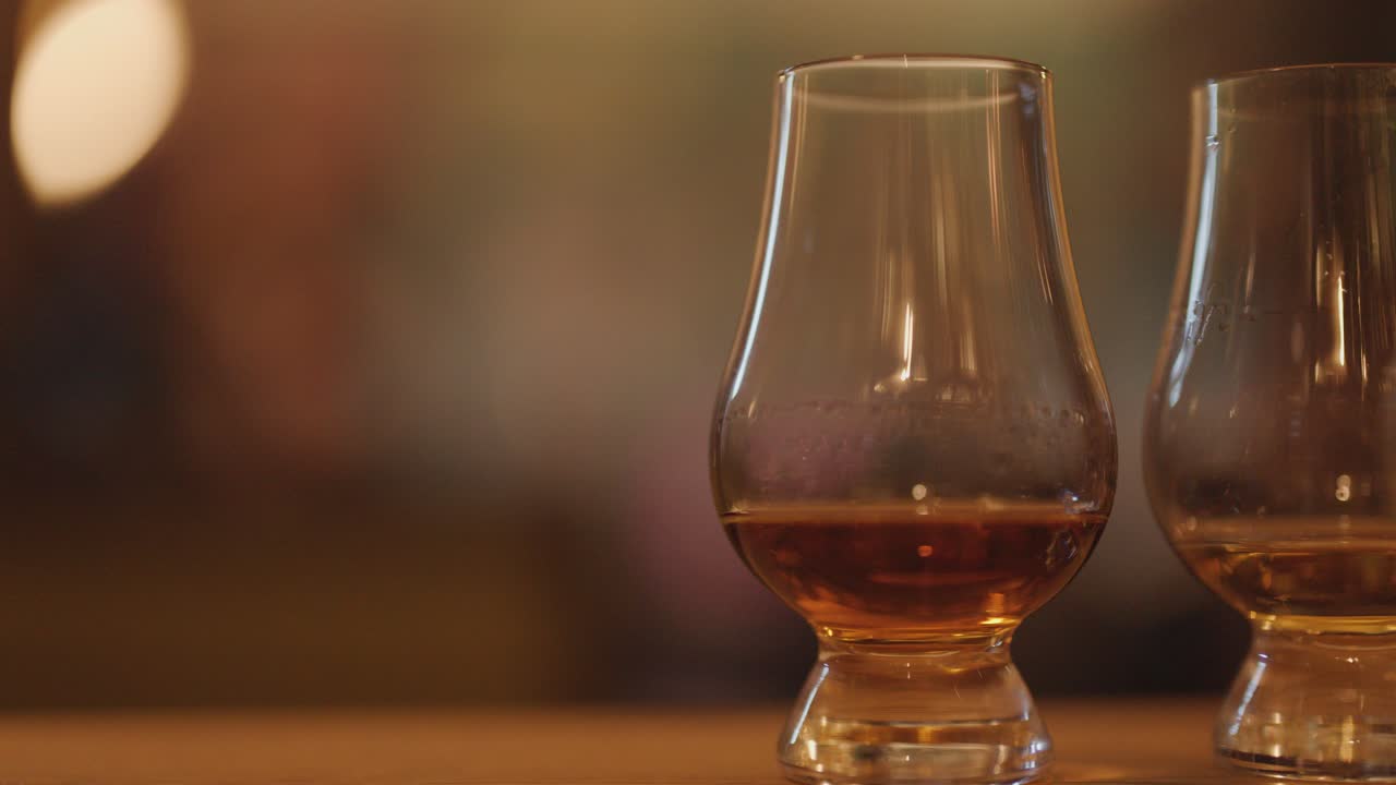 Two whisky glasses rest on a wooden bar, illuminated by soft, warm ambient lighting with a blurred background and shallow depth of field