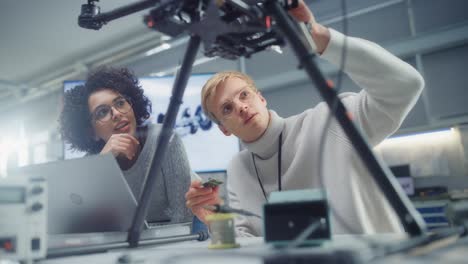 professional young black developer coding at laptop while her male colleague holding semiconductor at factory workshop. innovative technologies in unmanned aerial vehicle design concept