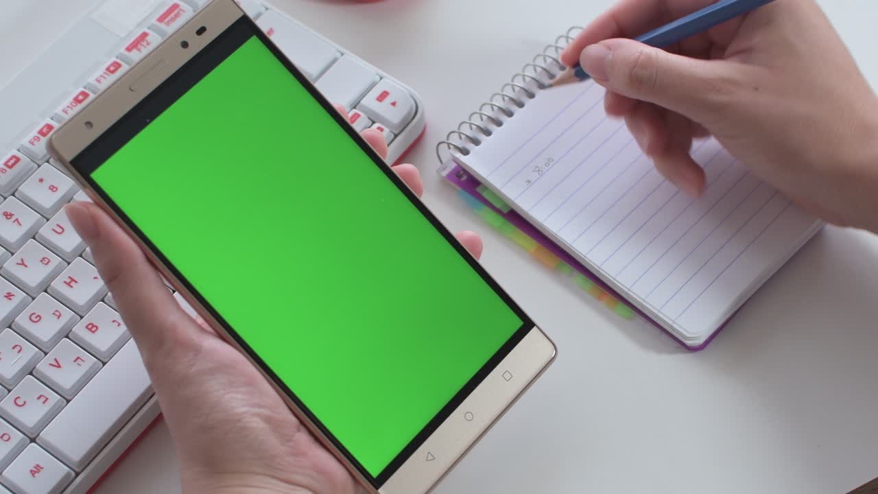 woman's hands holding a mobile with green screen, while taking notes in a notebook. Keyboard and white desktop.