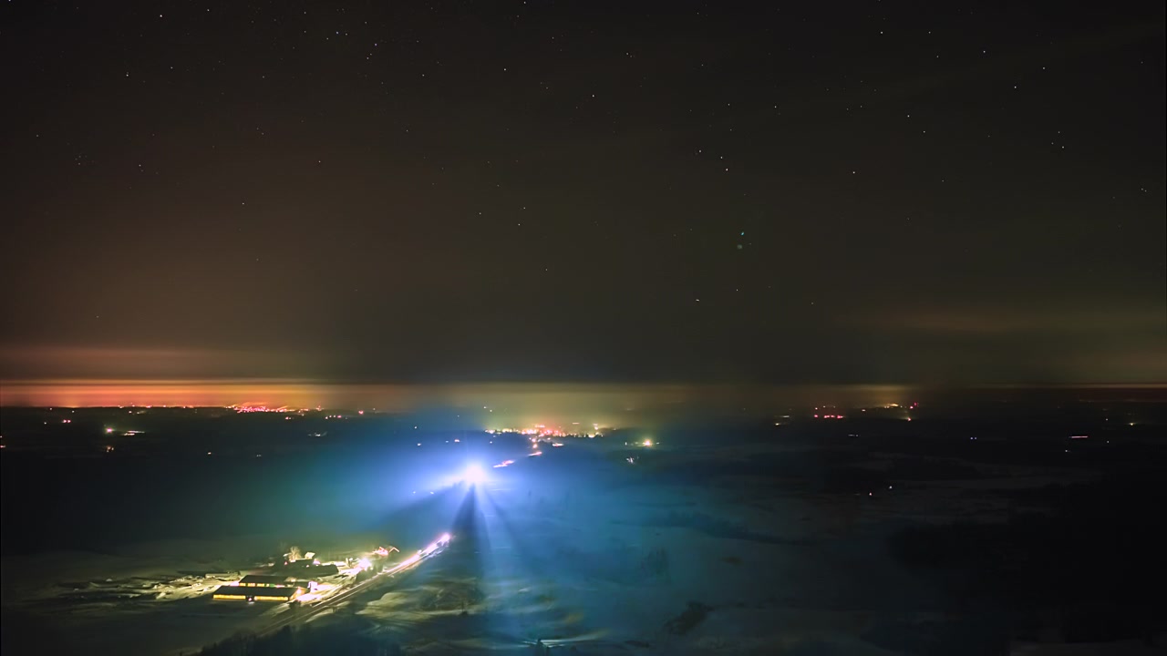 Uma paisagem noturna escura brilha com luzes distantes da cidade sob um céu estrelado e fracos feixes de luz verticais no horizonte - Timelapse.