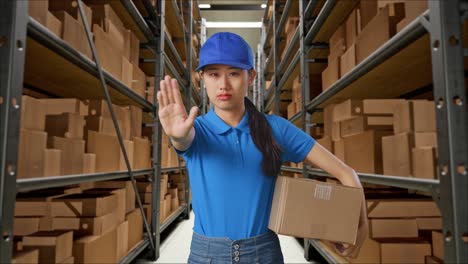 asian female courier in blue uniform making stop gesture and shaking her head while delivering a carton in warehouse