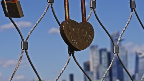A heart-shaped love lock engraved with “Love You Forever” hangs on a fence, with the New York City skyline softly blurred in the background