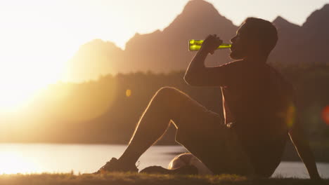 Hombre-Relajante-Sentado-Junto-Al-Lago-Y-Las-Montañas-Al-Atardecer-Bebiendo-Cerveza