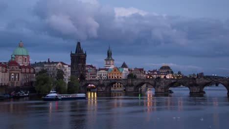 day to night sunset timelapse with a close up view of charles bridge on vltava river with boats on a cloudy evening in prague, czech republic as the bridge and towers of the city light up