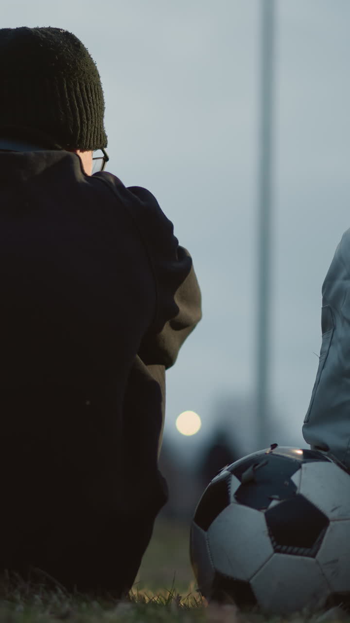 Two boys are seated closely together on a grassy field at dusk, with one resting his hand on a soccer ball, with a blur view of someone passing by in the background