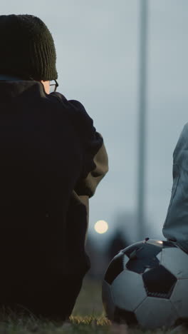 two boys are seated closely together on a grassy field at dusk, with one resting his hand on a soccer ball, with a blur view of someone passing by in the background
