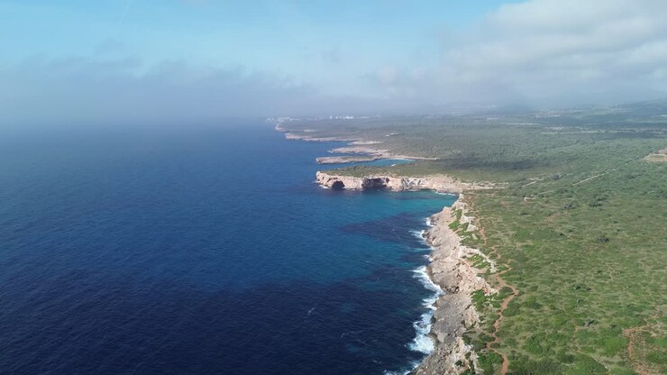 Ampia ripresa drone 4K della costa di Maiorca con il mare blu a sinistra e colline verdi lussureggianti a destra, sotto un cielo soleggiato con nuvole basse sparse. Catturato a giugno 2025