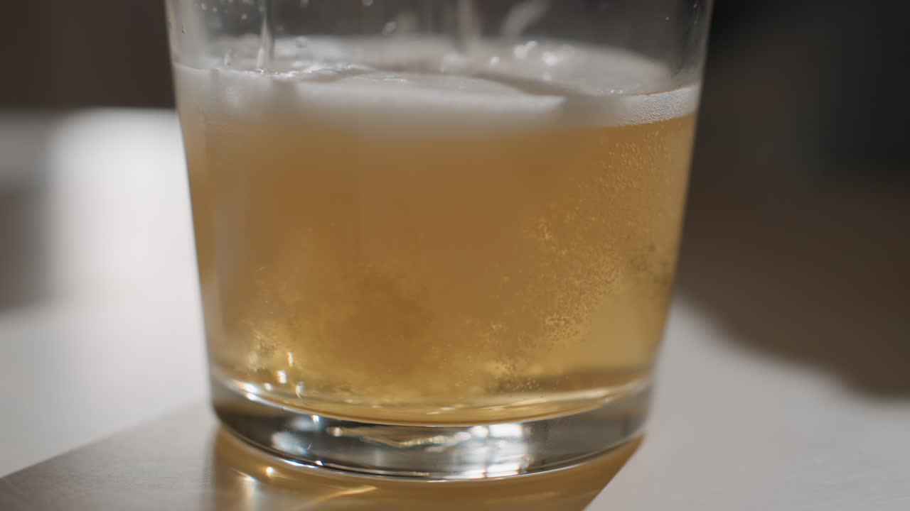 Primo piano di bicchiere di birra sul tavolo che mostra schiuma e microbolle che salgono, effervescenza delicata e formazione della schiuma, luce ambientale soffusa, degustazione in birrificio o controllo qualità del bar, consistenza cremosa e riflessi luminosi.