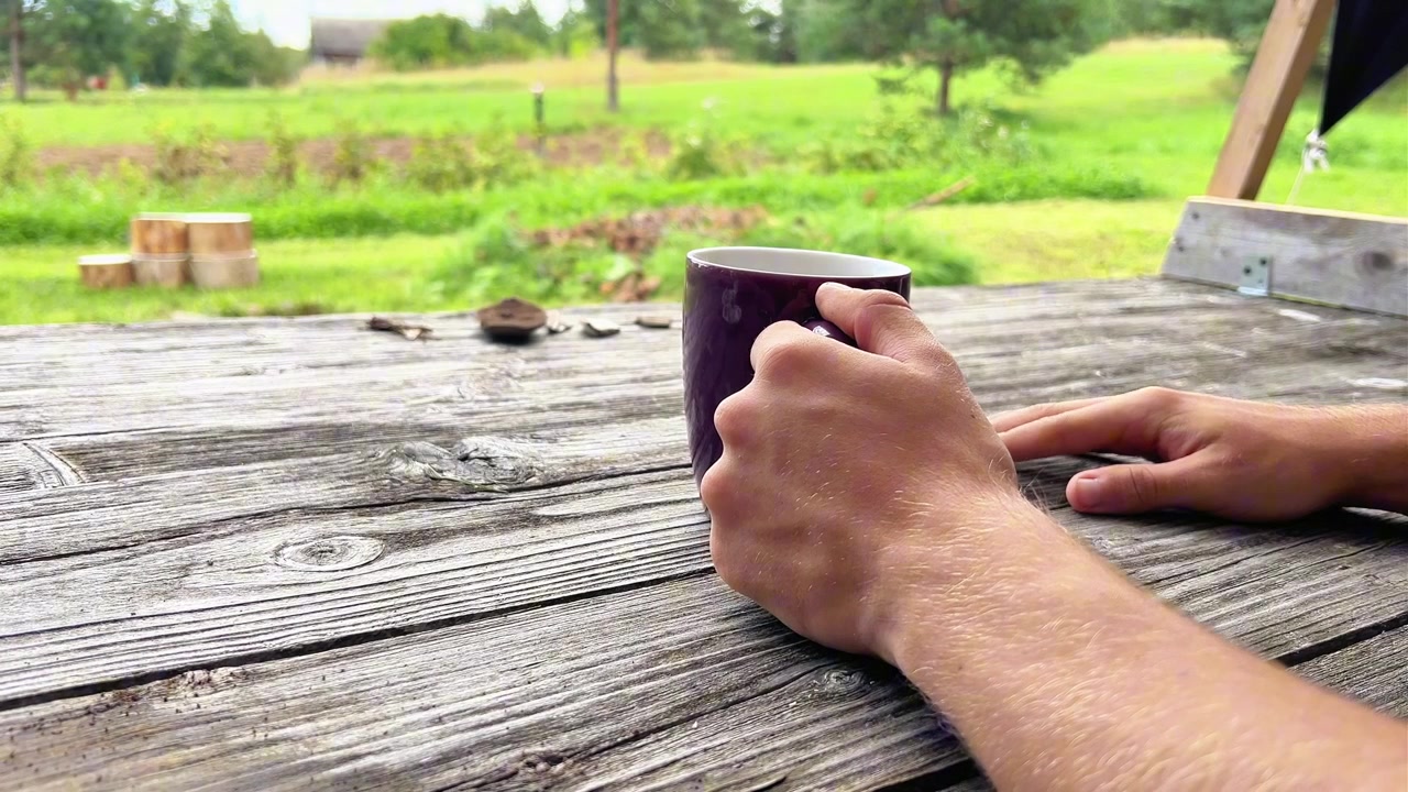 Close-up shot of a person holding a purple ceramic mug on a weathered wooden table with a rural backyard background. Ideal for morning routine ads, beverage promos, and lifestyle media.