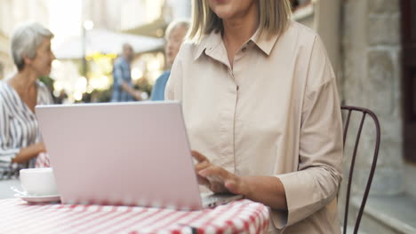 Ältere-Hübsche-Frau-Mit-Grauen-Haaren,-Die-Am-Tisch-Auf-Der-Caféterrasse-Sitzt-Und-Am-Laptop-Arbeitet