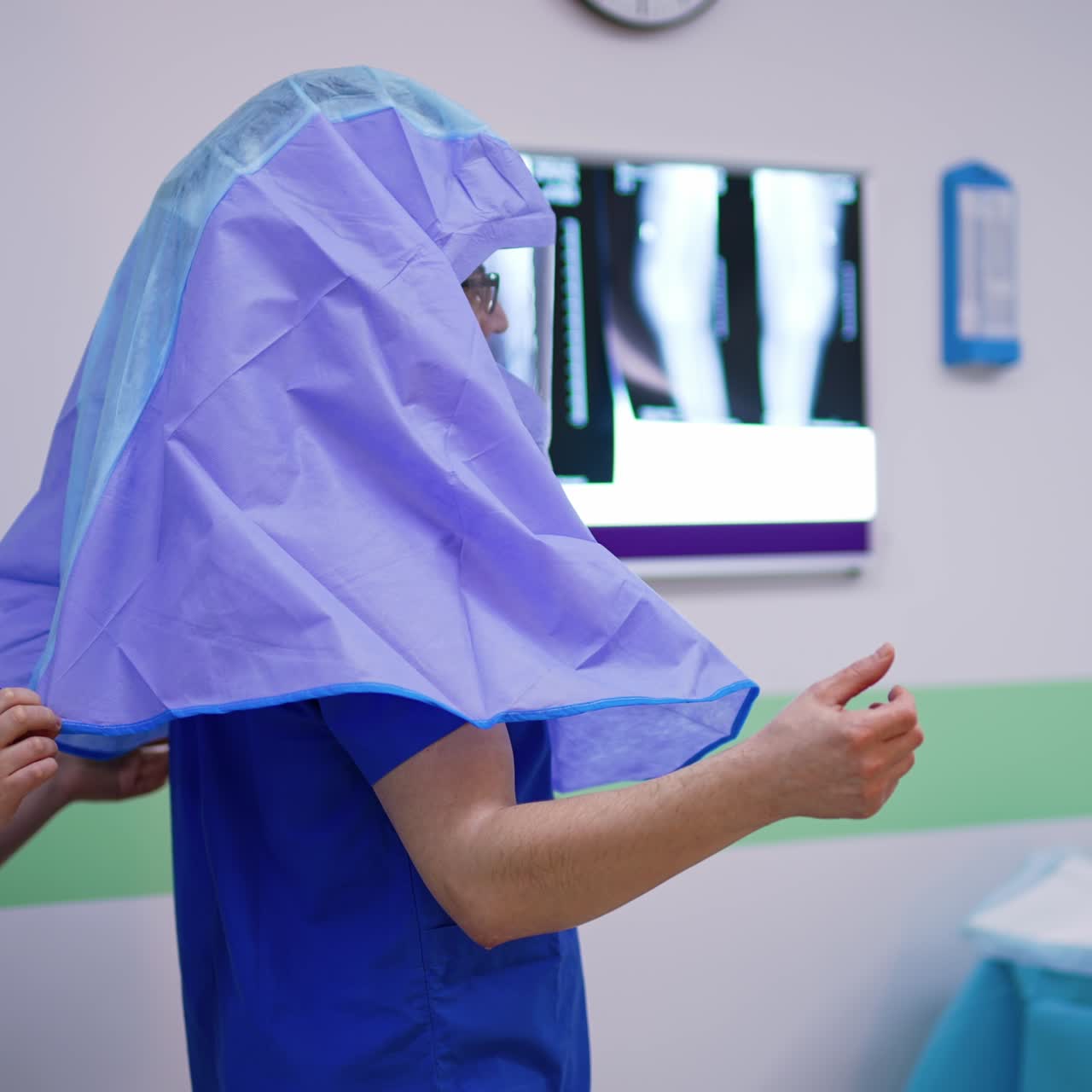 Male surgeon with helmet on his head stands holding clean hands to the sides. Female medical staff helping doctor to put on protective robe