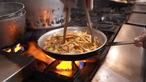 chef uses tongs to flip and toss traditional udon noodles with shrimp and vegetables in steel pan over fiery commercial stove, slow motion hd