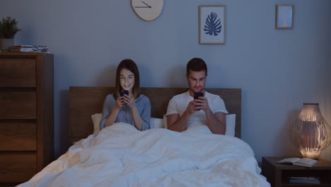 caucasian couple using smartphones while they are sitting in the bed under the blanket