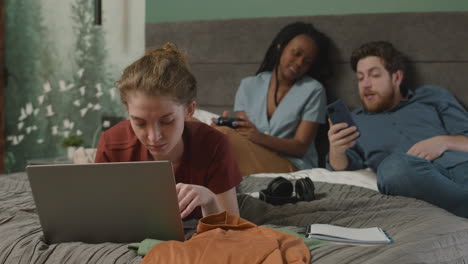 girl using laptop lying on the bed while her roommates are talking in the background