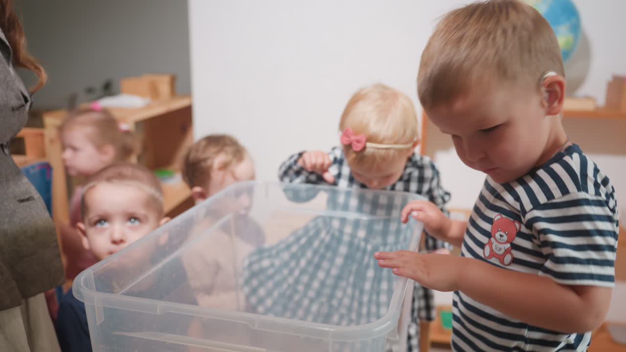 Toddlers gather around transparent container during classroom activity, one child in striped shirt with hearing aid smiling excitedly while teacher and peers watch with curiosity
