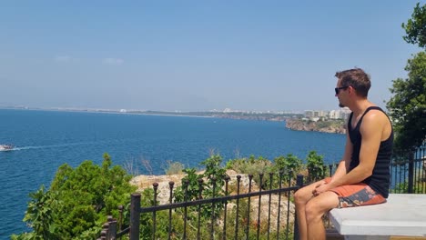 man in summer clothes and sunglasses smokes a cigarette above antalya’s shoreline, enjoying the wide panoramic view over the mediterranean on a windy spring day in turkey