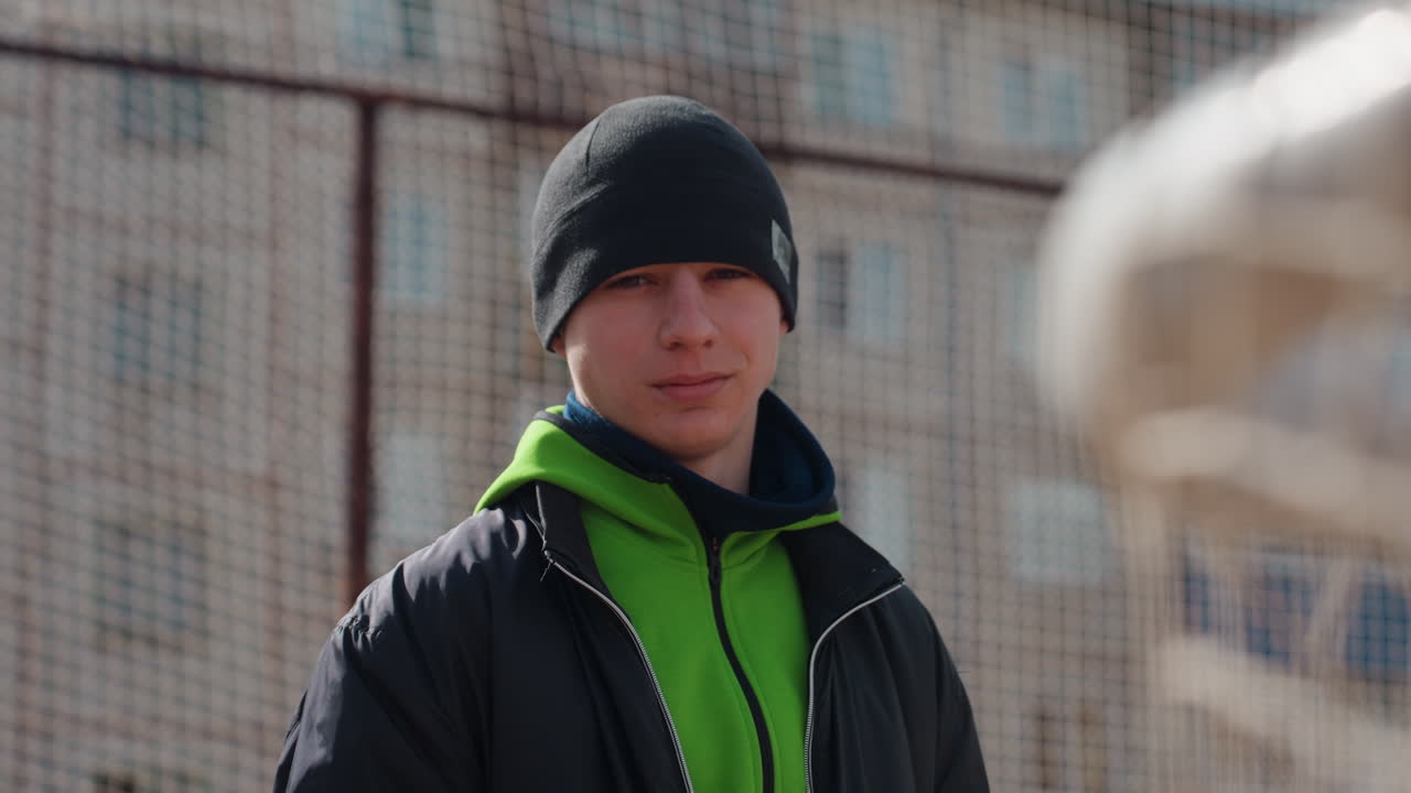 도시 코트에서 축구공을 들고 있는 백인 십 대; 사색에 잠긴 표정, 네온 후드티와 비니, 철조망 울타리와 아파트 블록 근처에 기대어 패스를 기다리는 모습, 차가운 공기 속에서 희미하게 보이는 입김