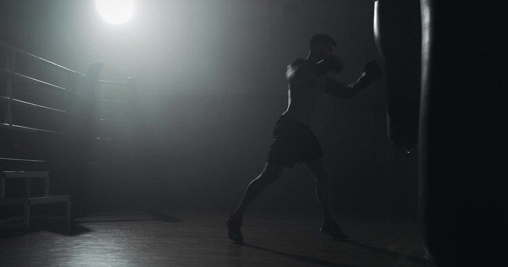 Silhouette di pugile che si allena con il sacco da boxe in una palestra buia.