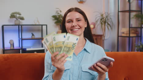 smiling happy rich young woman counting money euro cash use smartphone income saves lottery win