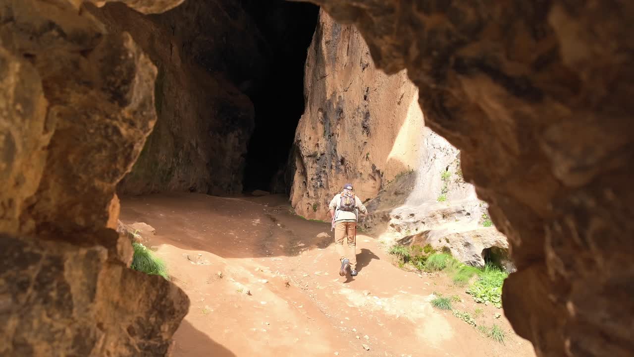 hombre blanco con una mochila y un bastón para caminar entrando en una caverna siniestra disparado por la espalda