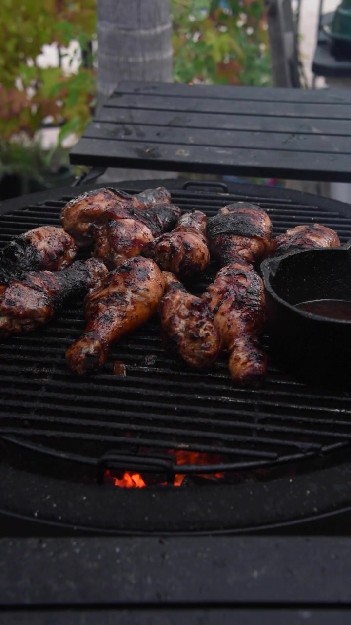 Video verticale in primo piano della mano di un uomo che usa delle pinze per girare cosce di pollo marinate. La carne sta cuocendo su un barbecue a carbonella caldo in un cortile.