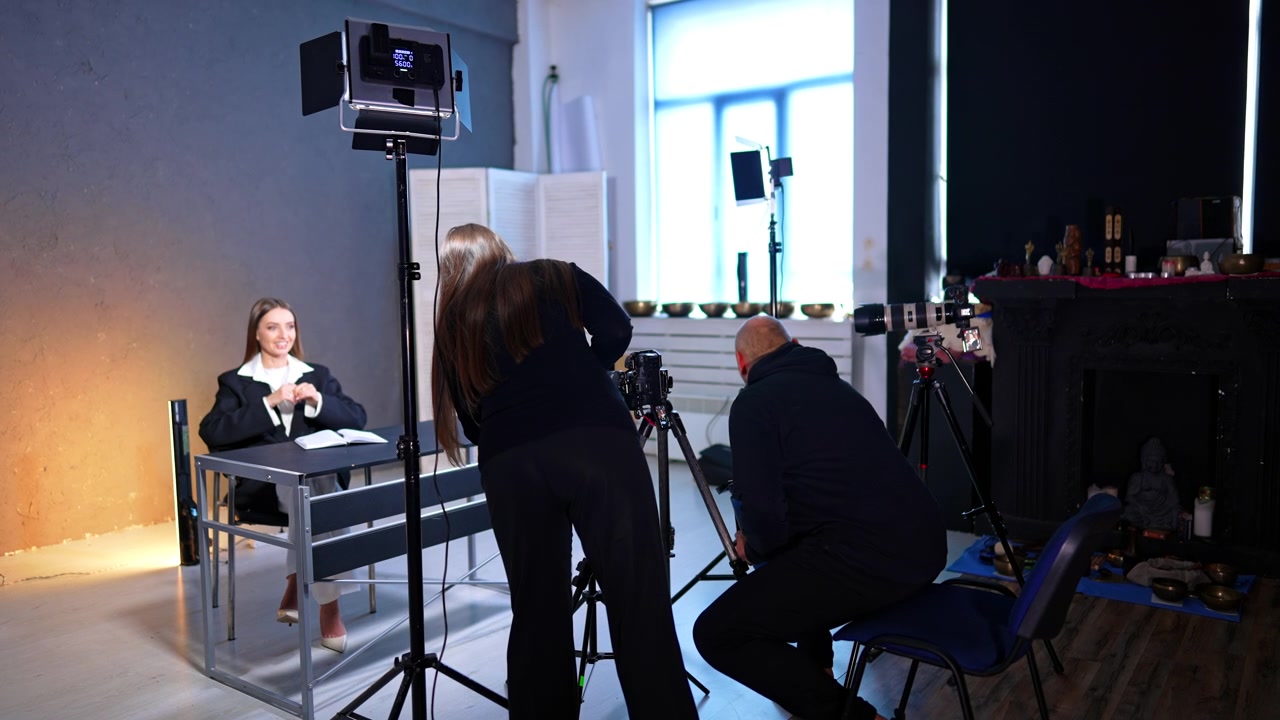 Kompletní studiové uspořádání s moderátorkou a štábem během video produkce. Široký záběr ukazující moderátorku u stolu a štáb s profesionálním osvětlením a kamerovou technikou.