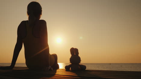 Eine-Frau-Sitzt-Auf-Einem-Steg-Neben-Einem-Spielzeughasen-Zusammen-Treffen-Sie-Den-Sonnenaufgang-über-Dem-Meer-Romantische-Stimmung