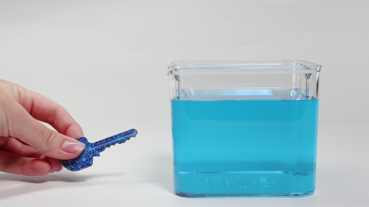 A hand places a textured key into a water-filled container, observing its buoyancy and sinking behavior