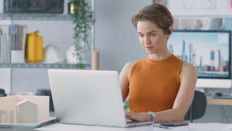 Female-Architect-In-Office-Listening-To-Music-On-Wireless-Earphones-Whilst-Using-Laptop