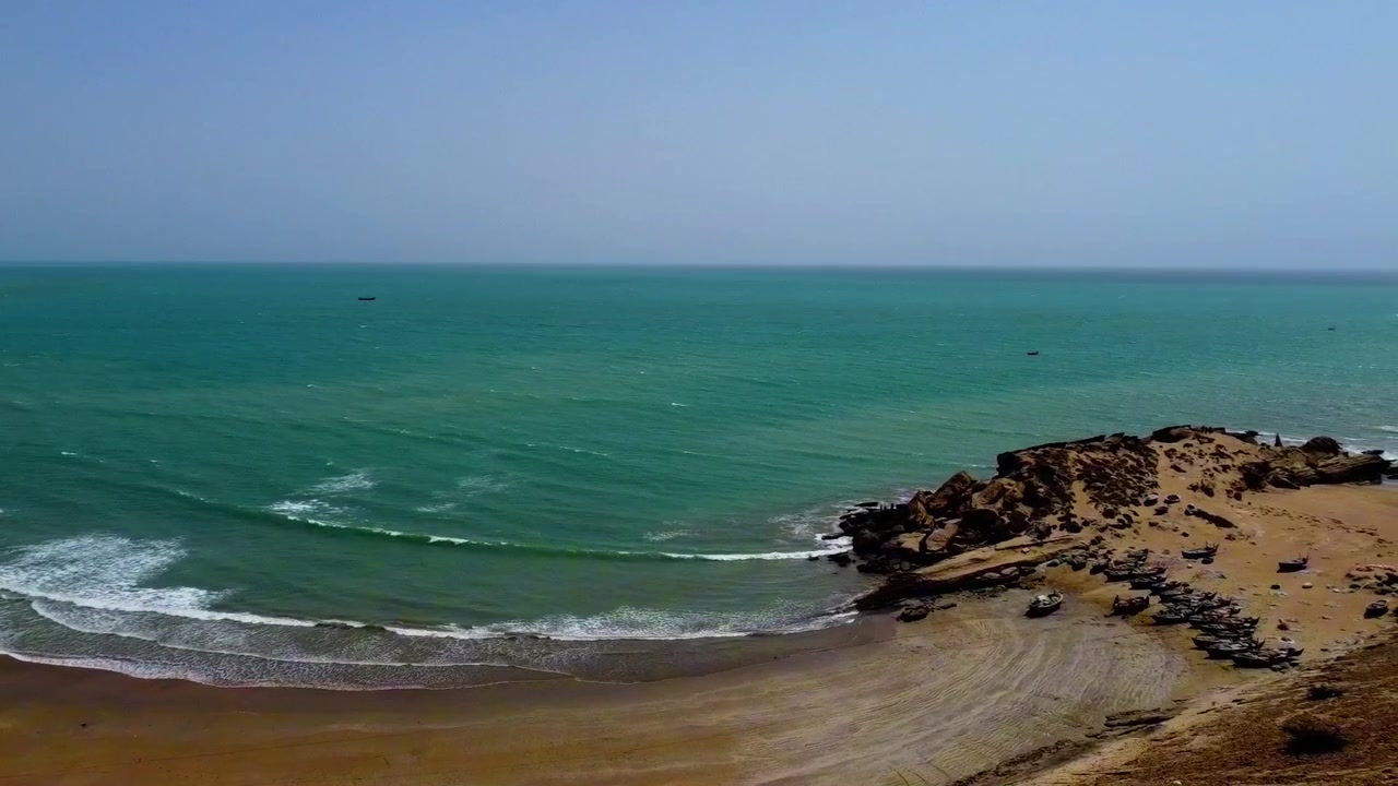 Video drone cattura un ampio panorama aereo della spiaggia di Kund Malir con acque turchesi, sabbia dorata, barche tradizionali in legno e pescatori che preparano le loro barche sotto cieli sereni e soleggiati