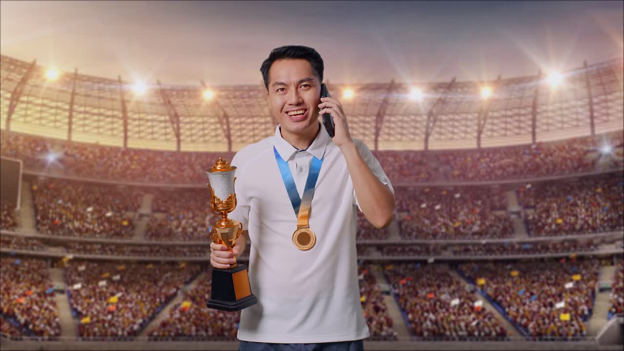 금메달을  아시아인 남자가 금 트로피를 들고 경기장에서 스마트폰으로 이야기하고 있다.