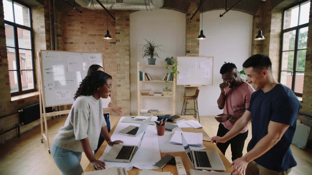 Gathering coworkers leaning over wooden table in loft office, consulting laptops, finalizing plan