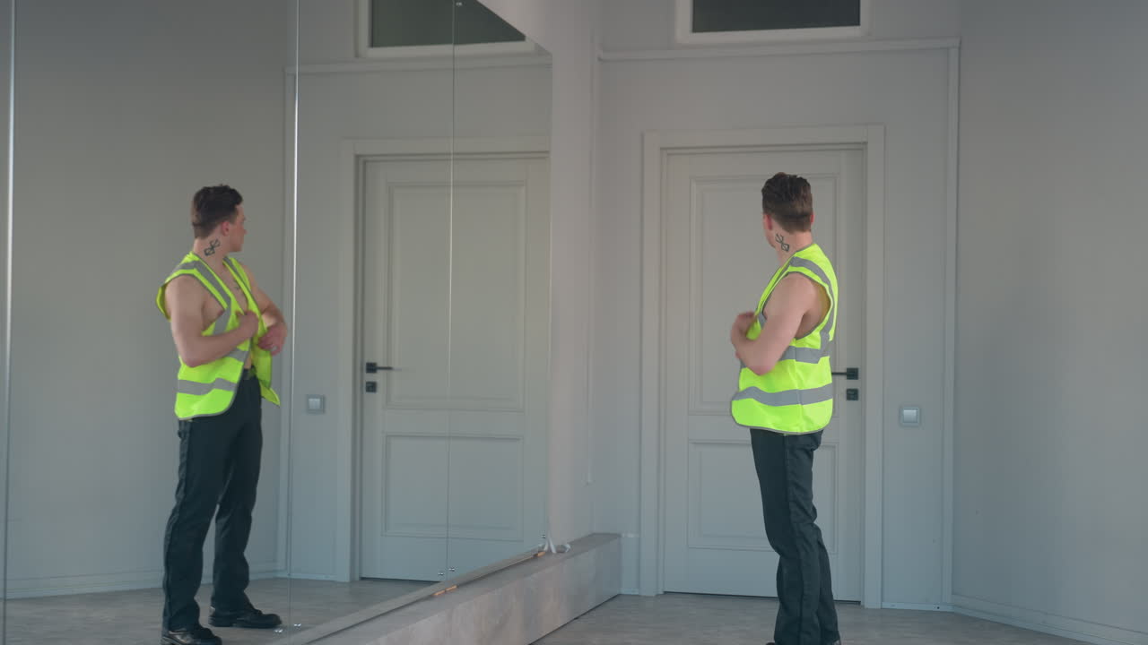 shirtless young man stands before full length mirror and closed door, pulling on neon yellow jacket with confident pose, showcasing bold neck and chest tattoos in dance studio with minimal decor