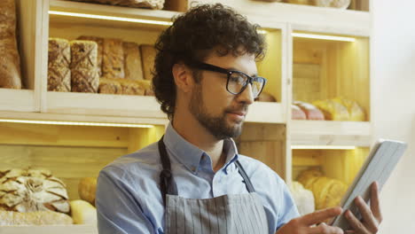 Handsome-Man-Bread-Seller-In-Glasses-Using-His-Tablet-Computer-While-Standing-At-The-Counter-In-The-Bakery-Shop