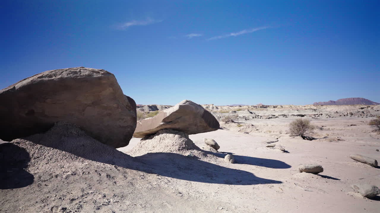 Slow right pan reveals striking rock formations casting long, defined shadows across the desert floor under a bright sun