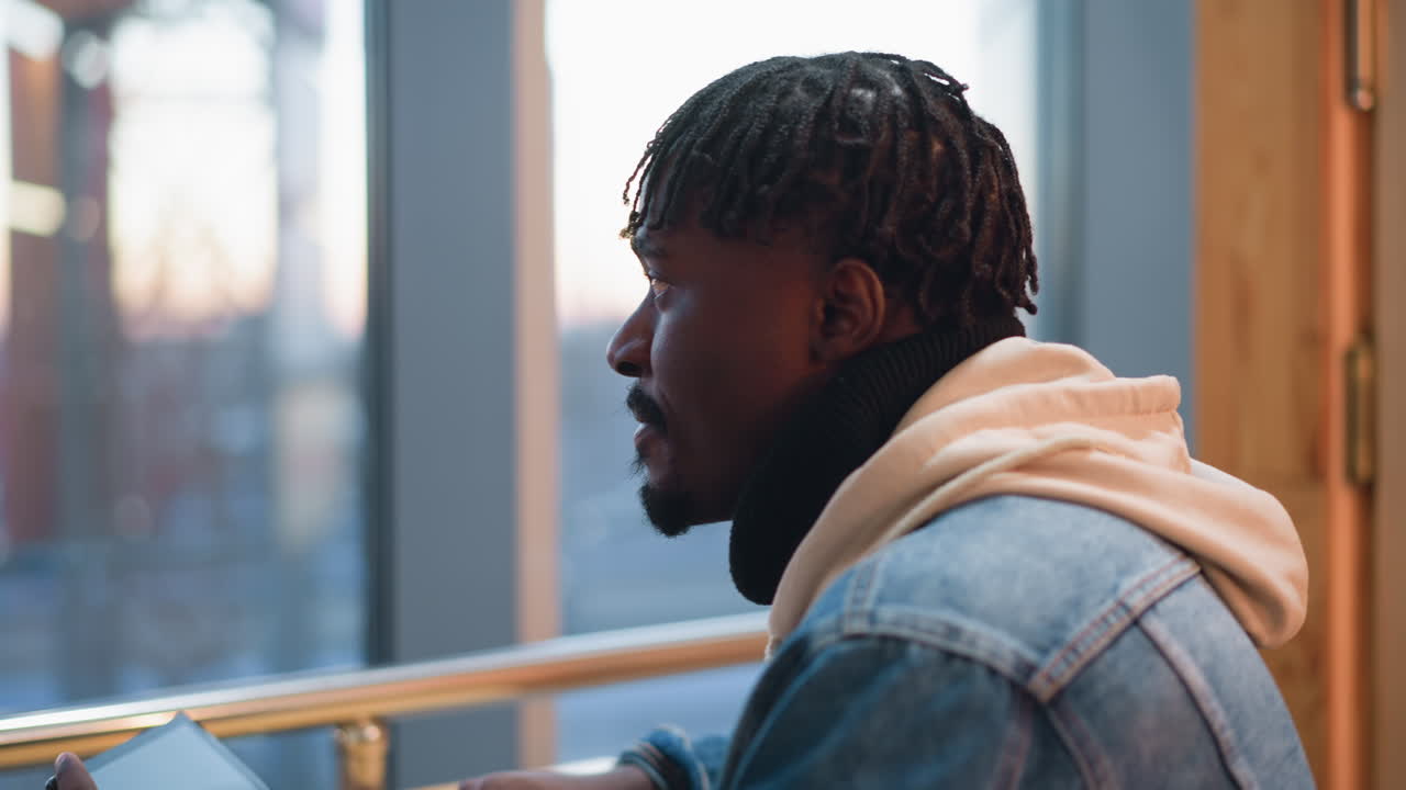 close up of young black man using smartphone while seated at wooden counter by large window with urban twilight skyline background wearing denim jacket and hoodie casual modern vibe