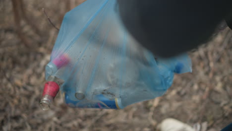 Volunteer wearing gloves and safety vest picking up dirty plastic bottle from ground covered with dry leaves during environmental cleanup, focusing on waste removal and pollution reduction