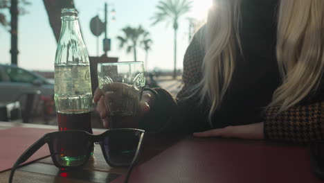 woman picks up glass from a cafe table where sunglasses and bottle stands lit by evening sunlight