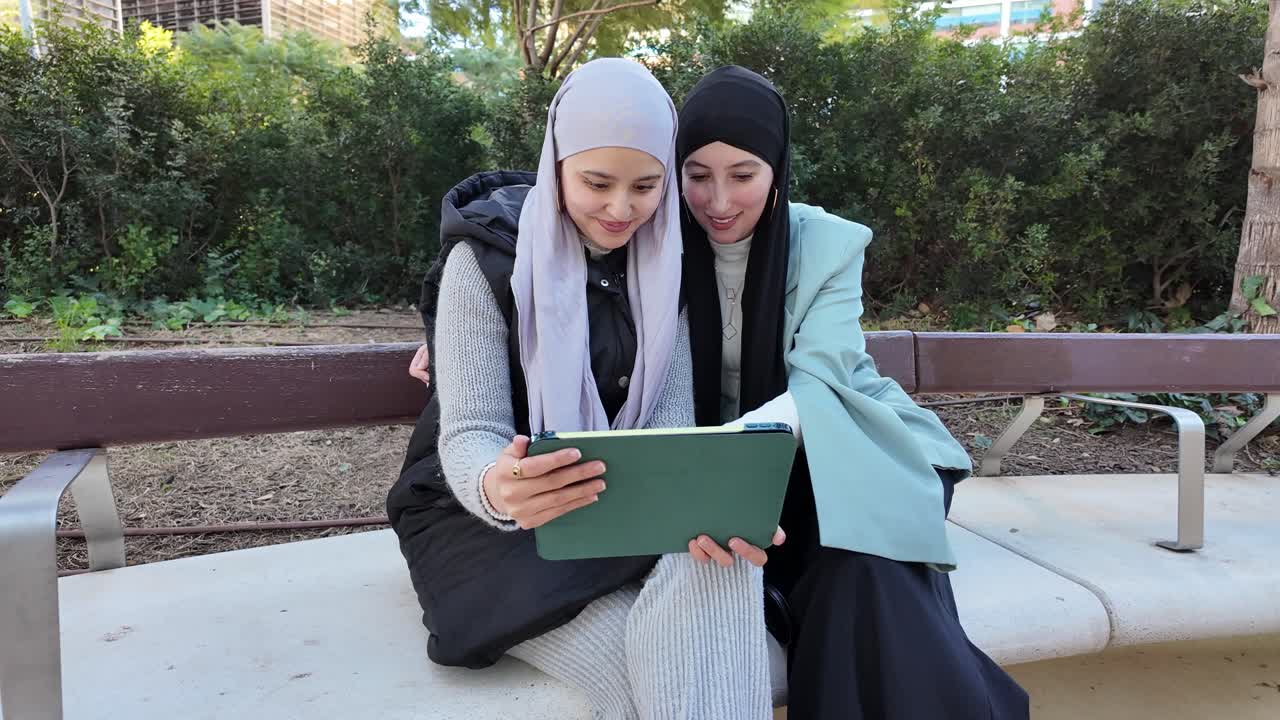 Two women with hijabs using a tablet on a bench in a park